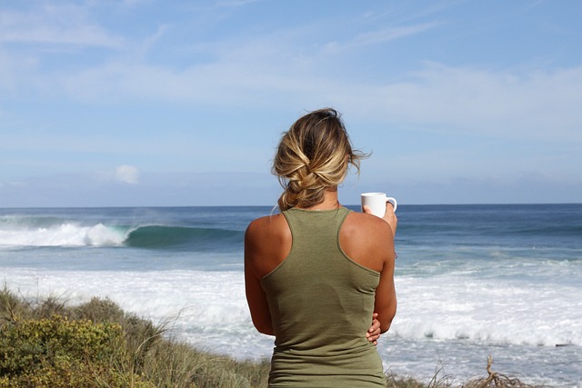 back of a woman drinking something out of a mug overlooking the ocean